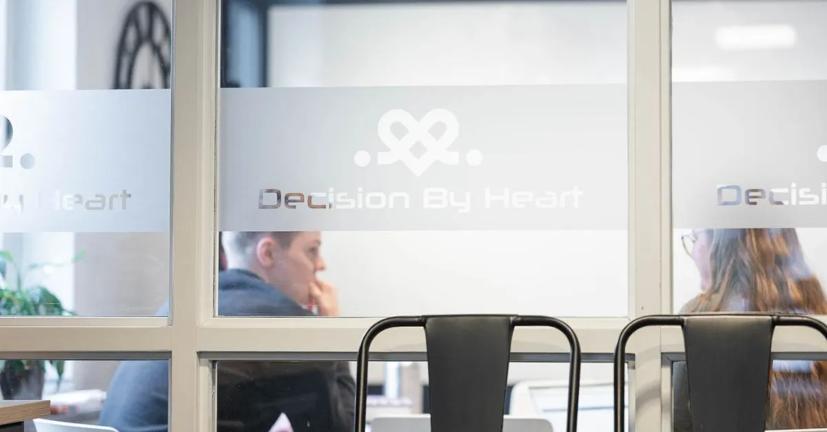 Två medarbetare på en webbyrå i Karlstad som diskuterar projekt genom en glasvägg med företagets logotyp 'Decision By Heart' tryckt på frostfilm.